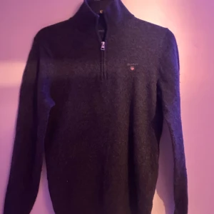 Mörkgrå tröja med dragkedja från GANT - Snygg mörkgrå tröja från GANT med halv dragkedja och broderad logotyp på bröstet. Perfekt för kyligare dagar med sin stilrena design och bekväma passform. Tveka inte om frågor!