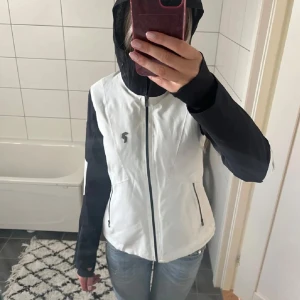 Vit och svart jacka från Peak Performance - Snygg vit och svart jacka från Peak Performance med huva och dragkedja. Jackan har en sportig look med kontrasterande svarta ärmar och detaljer. Perfekt för utomhusaktiviteter med praktiska fickor och en bekväm passform.