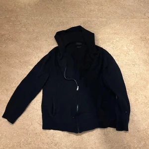 Svart merinojacka från Next Signature - Säljer en stilren svart hoodie från Next Signature i 100% merinoull. Jackan har en dragkedja framtill och långa ärmar. Perfekt för kyligare dagar med sin mjuka och varma känsla. Säljer på grund av att den har krympt i tvätten. Den är XL men skulle nu passa någon som har M eller L. Användes en gång. Priset kan diskuteras då jag köpte den för 600 kr.