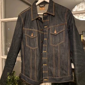Nudie Jeansjacka - Säljer denna svin goa jeansjacka/jeansskjorta från nudie jeans då den sitter för litet på mig. Den är knappt använd, i princip nyskick. Jackan är i dry/raw denim vilket innebär att materialet är helt obehandlat, detta gör delvis att det är ”rigid” vilket gör en outfit riktigt fet men också att du kommer skapa dina egna slitningar när du har på dig den, som på jeansbyxor. Denna modellen, Billy, är sitter mer slim jämfört med nudies andra jackor, så passar S-M! Skriv för frågor och prisförslag! 