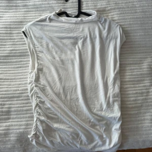 Vit ärmlös topp från Stradivarius - Säljer en stilren vit ärmlös topp från Stradivarius. Toppen har en snygg rynkad detalj på sidan som ger en unik touch. Perfekt för varma dagar eller som ett lager under en jacka.