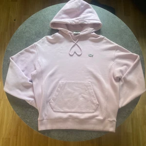 Ljusrosa hoodie från Lacoste - Säljer en snygg ljusrosa hoodie från Lacoste med klassisk krokodillogga på bröstet. Hoodien har en stor ficka framtill och en bekväm huva med dragsko. Perfekt för en avslappnad stil. Unisex-modell.
