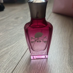 Rosa parfymflaska från Police - Snygg rosa parfymflaska från Police med en cool dödskalle-design på framsidan. Flaskan har en metallisk kork och en modern, kantig form som ger den ett edgy utseende. Perfekt för den som gillar en unik och iögonfallande stil.