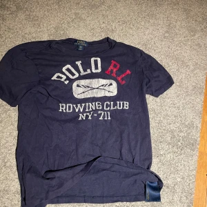 Blå t-shirt från Ralph Lauren - Säljer en blå t-shirt från Ralph Lauren med texten 'Polo RL Rowing Club NY-711' i vitt och rött på framsidan. T-shirten är kortärmad och tillverkad i bomull. Perfekt för en avslappnad stil.
