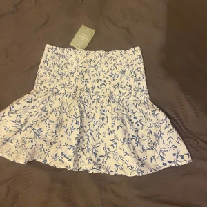 Vit blommig kjol - Charmig vit kortkjol med blått blommönster. Kjolen har en elastisk midja som ger en bekväm passform och en volangdetalj som ger extra stil. Perfekt för varma dagar! Pris kan diskuteras, skriv för fler bilder.