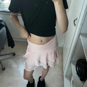 Ljusrosa volangkjol - Säljer en superfin ljusrosa kortkjol med volanger. Perfekt för sommardagar eller en festlig kväll. Kjolen har en elastisk midja som gör den bekväm och lätt att bära. Materialet är lätt och luftigt, vilket gör den idealisk för varma dagar. Passar perfekt med en enkel topp eller en snygg blus!