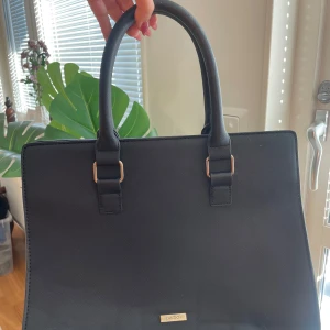 Svart handväska från Aldo - Säljer en stilren och elegant svart handväska från Aldo. Väskan har en klassisk rektangulär form med två robusta handtag och en praktisk dragkedja upptill. Perfekt för både vardag och mer formella tillfällen. Den är i mycket gott skick och redo att användas direkt!