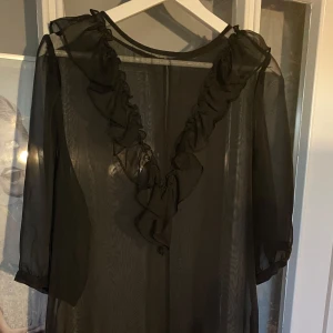 Svart transparent blus med volangdetaljer - Säljer en elegant svart transparent klänning med vackra volangdetaljer framtill. Klänningen har trekvartsärmar och en lös passform. 