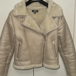 Beige jacka från Missguided - Säljer en superfin beige jacka från Missguided. Den har en snygg asymmetrisk dragkedja och två fickor med dragkedjor framtill. Jackan är fodrad med mjukt fuskpälsmaterial som gör den perfekt för kyliga dagar. Den har en croppad passform och är långärmad. Perfekt för höst och vinter!