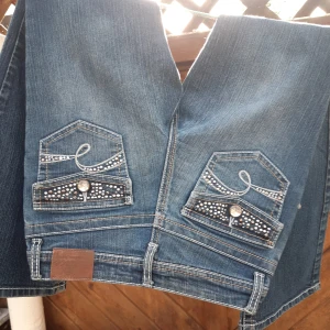 Blå jeans med strassdetaljer - Säljer ett par snygga blå jeans med strassdetaljer på bakfickorna och framfickorna. De har en bootcut-stil som ger en skön retrovibe. Perfekta för en festlig look eller bara för att liva upp vardagen! 💎 Midjemått 31cm innebenslägd 70 cm