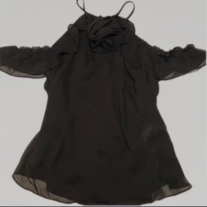 Svart topp - Snygg svart topp med volangdetaljer vid axlarna och en blomdetalj vid halsen. Toppen är i en lätt och luftig kvalitet. Stängda med knapp i nacken. Är i storlek L men passar från M-L, S om du vill ha den lite lösare. 