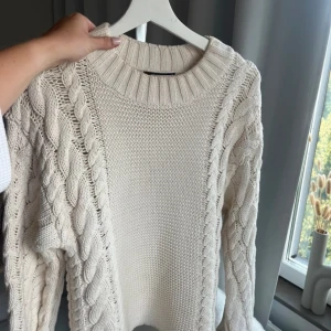 Vit stickad tröja med flätmönster - Säljer en supermysig vit stickad tröja med snyggt flätmönster på ärmarna och sidorna. Perfekt för kyliga höst- och vinterdagar. Tröjan har en rund halsringning och långa ärmar. Den är både stilren och bekväm, perfekt för att mysa i hemma eller för en dag ute i stan. 🧶✨