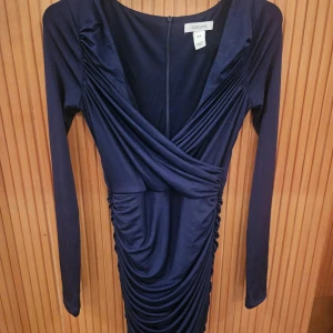 Mörkblå omlottklänning från Oneness - Säljer en elegant mörkblå omlottklänning från Oneness i storlek XS. Klänningen har långa ärmar och en smickrande omlottdesign med draperade detaljer. Perfekt för en festlig kväll eller en speciell tillställning.