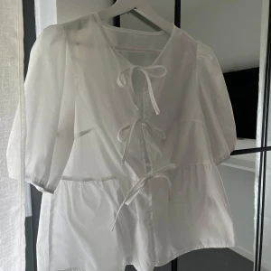 Vit blus med knytning - Säljer en superfin vit blus med knytning framtill. Den har puffiga trekvartsärmar och en luftig passform. Blusen är i ett lätt och bekvämt material, vilket gör den perfekt för både vardag och fest. 