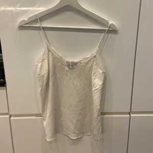 Vit sidenlinne med spetsdetaljer - Säljer ett elegant vitt sidenlinne med tunna axelband och vacker spetsdetalj längs kanten. Perfekt för sommardagar eller som ett lager under en kavaj. Linnet har en liten defekt som syns på bilden, men det påverkar inte användningen. 🪽