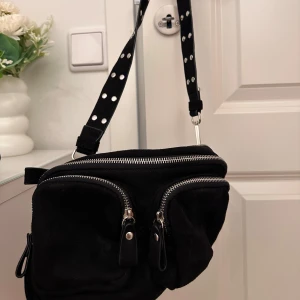 Svart axelväska med dragkedjor - Säljer en supercool svart axelväska med silverfärgade detaljer. Väskan har massa praktiska fickor och rymmer definitivt mer än vad man kan tro. Använd väldigt sparsamt!