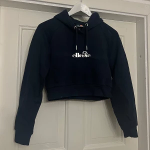 Mörkblå croppad hoodie från Ellesse - Marinblå croppad hoodie från Ellesse. Den har en klassisk huva med dragsko och ett tryck med Ellesse-loggan på bröstet. Storlek 36. Fint skick. 
