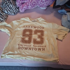 Gul t-shirt från H&M med tryck - Säljer en gul t-shirt från H&M med coolt tryck på framsidan. Texten 'LAKEWOOD 93 ROSE HILLS PALMDALE DOWNTOWN' ger en skön retrovibe. Perfekt för en avslappnad stil. T-shirten är kortärmad och i bomull, vilket gör den superbekväm att ha på sig. Passar perfekt till jeans eller shorts!