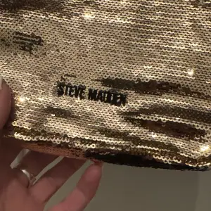 Snygg axelväska från Steve Madden i glittrande guld med paljetter. Väskan har en kedjerem och dragkedja upptill, perfekt för att lysa upp din outfit.