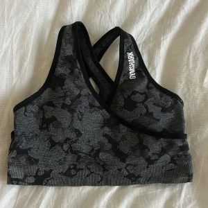 Svart och grå sporttopp från Gymshark - Snygg och bekväm sporttopp från Gymshark i svart och grått med ett subtilt mönster. Toppen har en korsad ryggdesign för extra stöd och komfort under träning. Storlek M. 