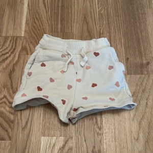 Vita shorts med hjärtan från Sweetie - Söta vita shorts från Sweetie med rosa och röda hjärtan. De har en bekväm resårmidja och snörning framtill. Perfekta för varma sommardagar! 💖