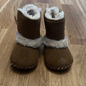 Bruna babyboots med pälsfoder - Söta bruna babyboots med mjukt vitt pälsfoder och dekorativ knappdetalj på sidan. Perfekta för att hålla små fötter varma under kyliga dagar. Sulan har blå detaljer för extra grepp.