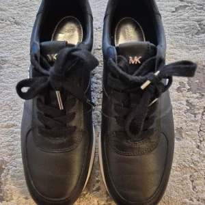 Svarta sneakers från Michael Kors - Snygga svarta sneakers från Michael Kors i skinn med silverdetaljer. De har en bekväm passform och snörning framtill. Perfekta för både vardag och festliga tillfällen. Sulan är vit med en cool design som ger extra stilpoäng.