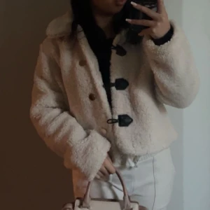 Beige teddyjacka med knappar - Mysig beige teddyjacka med stora knappar och en croppad passform. Perfekt för kyliga dagar när du vill vara både varm och stilren. Jackan har långa ärmar och en mjuk, fluffig yta.