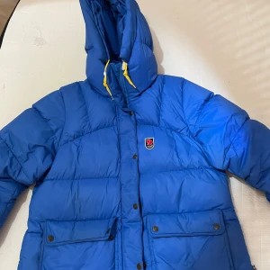 Blå dunjacka från Fjällräven - Snygg blå dunjacka från Fjällräven med huva och gula detaljer. Jackan har en klassisk design med knappar och dragkedja framtill samt praktiska fickor. Perfekt för kyliga dagar. Storlek M 