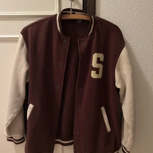 Brun och vit jacka från H&M - Snygg brun och vit jacka från H&M med bokstaven 'S' på framsidan och texten 'Seattle' på baksidan. Jackan har långa ärmar och stängs med knappar. Perfekt för en avslappnad stil.