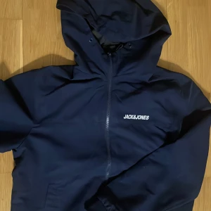 Mörkblå regnjacka från Jack & Jones - Säljer en snygg mörkblå regnjacka från Jack & Jones i storlek S. Den har en praktisk dragkedja och en huva för extra skydd. Perfekt för höst och vår! Jackan är i mycket bra skick och redo för nya äventyr.