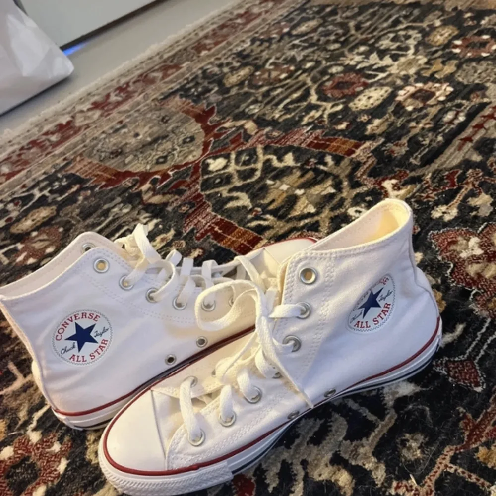 Säljer ett par klassiska vita Converse All Star sneakers med hög snörning och röd/blå randdetalj på sulan. Perfekta för en avslappnad stil. Aldrig använda endast testade💖. Skor.