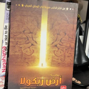 أرض زيكولا - En fascinerande roman med ett mystiskt omslag som visar en ljusstråle som bryter genom en stenmur. Boken är skriven på arabiska och verkar vara en populär titel i arabvärlden. Perfekt för dig som älskar äventyr och mystik.