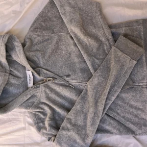 Grå hoodie från Juicy Couture - Säljer en stilren grå hoodie från Juicy Couture med dragkedja och huva. Tröjan har långa ärmar och en liten broderad logga på bröstet. Perfekt för en avslappnad look.