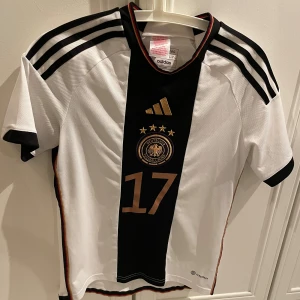 Tyskland match tröja från säsong 22/23 - Säljer en vit Adidas fotbollströja med svarta och guldiga detaljer. Tröjan har nummer 17 och namnet Wirtz på ryggen. Den är kortärmad och i mycket bra skick. Perfekt för unga fotbollsfans!