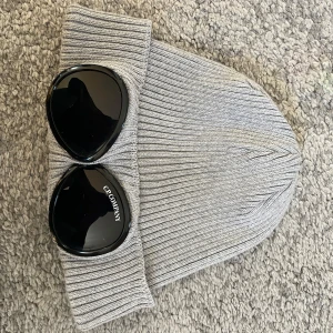 Grå ribbad mössa från C.P. Company - Snygg grå ribbad mössa från C.P. Company med coola glasögondetaljer på uppviket. Inte använd mer än ett fåtal gånger köpt hos miinto kvitto finns. 