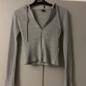Grå ribbad kofta från Gina Tricot - Säljer en snygg grå ribbad kofta från Gina Tricot i storlek XS. Den har en dragkedja framtill och långa ärmar, perfekt för lager på lager-stil. Koftan är croppad och har en huva med snören. Perfekt för både vardag och mysiga kvällar!