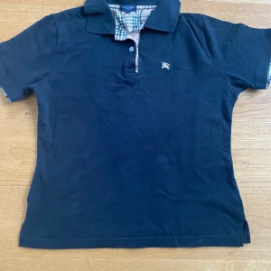 Svart kortärmad pikétröja från Burberry - Snygg svart pikétröja från Burberry med klassisk krage och korta ärmar. Tröjan har ett diskret broderat emblem på bröstet och en stilren rutmönstrad insida av kragen. Perfekt för en avslappnad men stilfull look.