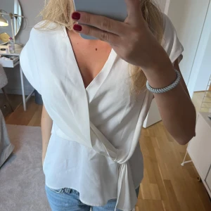 Vit omlottblus från Zara - Säljer en  vit omlottblus från Zara. Blusen har korta ärmar och en knytning i sidan. Aldrig använd! 