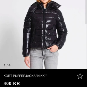 Kort svart pufferjacka 'Nikki' den blå knappt använd - Snygg och trendig kort svart pufferjacka med glansig yta och dragkedja framtill. Perfekt för kyliga dagar med sin vadderade design och höga krage.