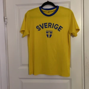 Gul Sverige t-shirt med blå detaljer - Snygg gul t-shirt med blå text 'Sverige' och ett svenskt flaggmärke på bröstet. Perfekt för att visa ditt stöd för Sverige! Kortärmad och bekväm passform. Storlek S