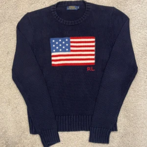 Marinblå Kabelstickad Tröja från Ralph Lauren med Amerikanska Flaggan - Säljer en snygg mörkblå stickad tröja från Polo Ralph Lauren med en stor amerikansk flagga på framsidan. Tröjan har långa ärmar och ribbstickade muddar vid ärmslut och nederkant. Perfekt för en stilren look!