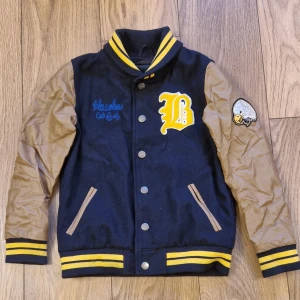 Svart och brun jacka från Soul & Glory - Snygg svart och brun jacka från Soul & Glory med broderade detaljer och knappar. Jackan har långa ärmar och ribbade muddar i svart och gult. Perfekt för en cool och avslappnad stil.