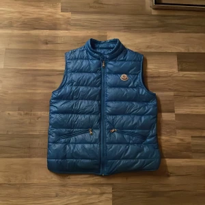 Blå dunväst från Moncler - Snygg blå dunväst från Moncler med dragkedja och två praktiska fickor framtill. Västen har en quiltad design och Moncler-logga på bröstet. Perfekt för kyligare dagar.