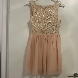 Beige paljettklänning - Snygg ärmlös klänning i beige med glittrig paljettöverdel och en luftig kjol. Klänningen har en öppen rygg med knytband för extra detalj. Perfekt för festliga tillfällen!