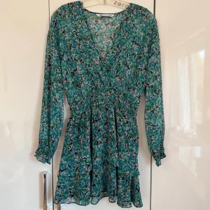 Blommig klänning från Zara - Säljer en superfin blommig klänning från Zara i storlek L. Den har en vacker grön och blå färg med blommönster och långa ärmar. Perfekt för vår och sommar! Klänningen har en smickrande passform med resår i midjan och en lätt volangdetalj vid fållen.