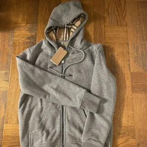 Grå hoodie från Burberry - Snygg grå hoodie från Burberry med dragkedja och klassiskt rutigt foder i huvan. Tröjan har långa ärmar och ribbade muddar. Perfekt för en stilren och bekväm look. Tröjan är använd endast engång tagsen finns kvar och medföljer. Pris kan diskuteras vid snabbaffär 