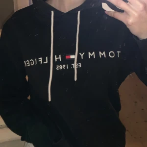 Svart hoodie från Tommy Hilfiger - Säljer en svart hoodie från Tommy Hilfiger med vit text och logga på framsidan. Den har en klassisk känguruficka och vita dragsnören vid huvan.