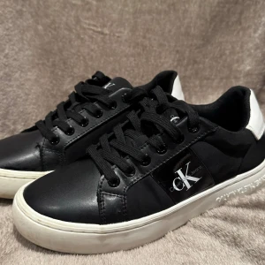 Svarta sneakers från Calvin Klein - Snygga svarta sneakers från Calvin Klein i syntetmaterial med vita detaljer. De har en klassisk snörning och en bekväm sula. Perfekta för vardagsbruk och ger en stilren look. Passar till både jeans och mer uppklädda outfits.
