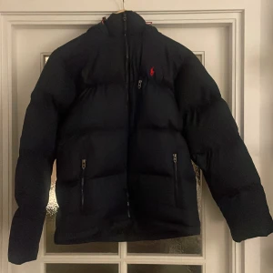 Mörkblå  dunjacka från Polo Ralph Lauren - Säljer en snygg mörkblå dunjacka från Polo Ralph Lauren, herr storlek Medium. Den har en klassisk design med dragkedja och det ikoniska röda logotypbroderiet på bröstet. Jackan är perfekt för kalla vinterdagar och har en skön passform med långa ärmar. Den är helt oanvänd. Säljes pga fel storlek.
